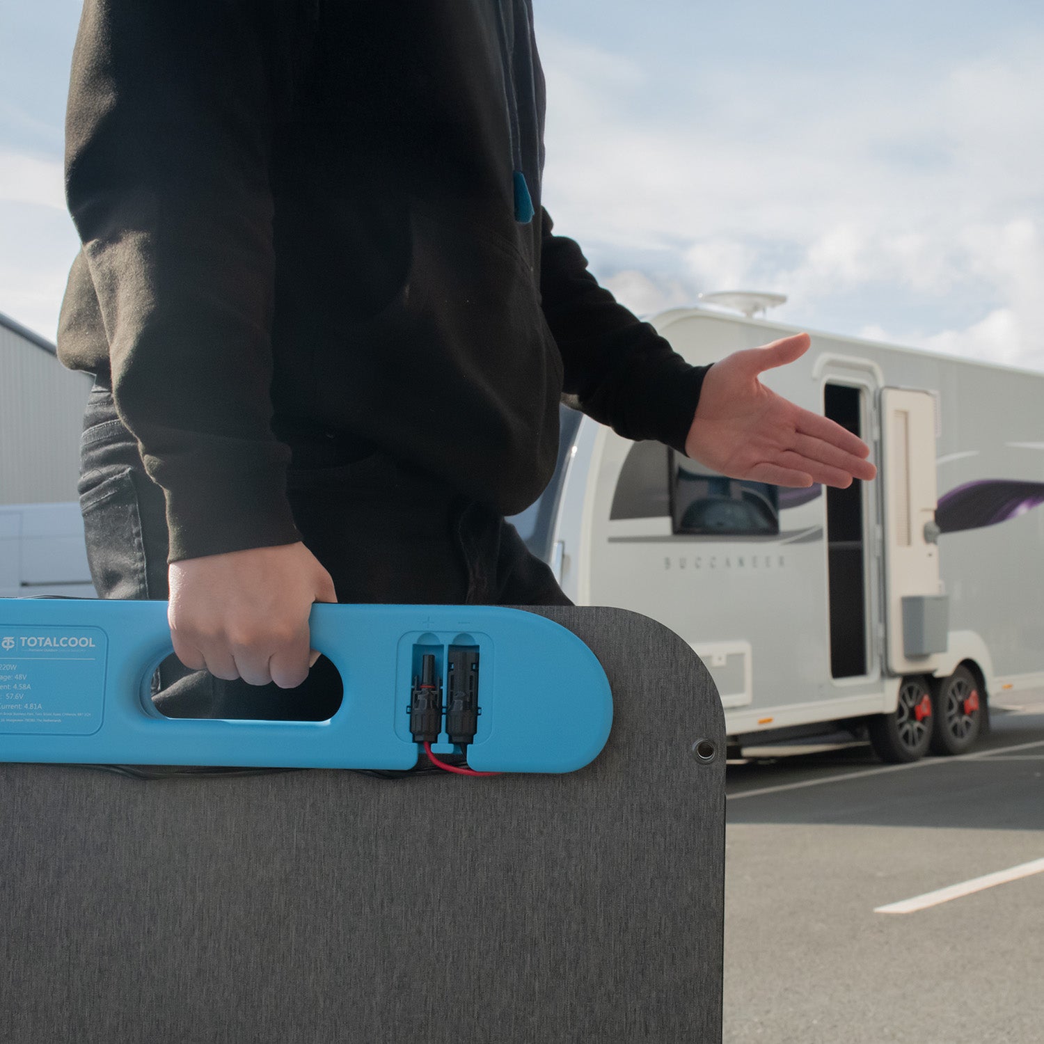 Person holding a blue and black foldable solar panel with a caravan in the background